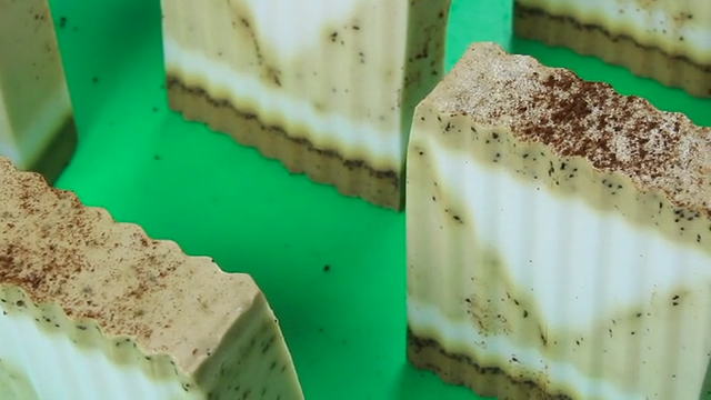 Sliced melt-and-pour soap bars with a cream layer between two coffee-flecked tan layers, dusted with coffee grounds on top, on a bright green surface
