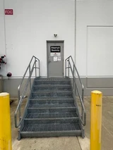 Metal stairs with handrails between yellow bollards leading to a gray pickup door