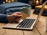 Hand holding a gold credit card over an open laptop with a mug and books on a wood desk