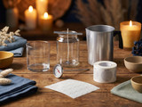 Candle-making supplies on a wood table: empty glass tumbler, wicked jar, dial thermometer, aluminum pouring pot, wick stickers, and a roll of caution labels, with lit candles glowing behind