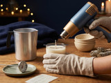 Gloved hand aiming a blue and gold heat gun over a wicked candle, beside an aluminum pouring pot, a dial thermometer on a plate, and a second work glove