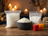 Bag of white wax pastilles and a bag of white powder flanking a dark bowl of soy wax flakes, with red and orange votives and lit candles behind, on a wood table