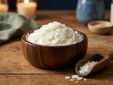 White soy wax flakes heaped in a wooden bowl with a wooden scoop on a rustic table, lit candles behind