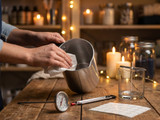 Hands wiping a stainless pouring pot with a paper towel beside a dial thermometer, clip thermometer, wicked jar, and lit candles