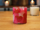 Glass candle vessel covered in overlapping red and pink tissue-paper hearts on a wood table beside a lit white candle