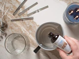 Overhead shot of a hand pouring a Lone Star fragrance oil bottle into a metal pour pot beside wicks, an empty jar, and a finished blue candle