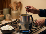 Hands checking a thermometer in a metal pouring pot set in a water bath over a gas flame, soy wax flakes in a bowl beside