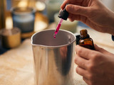 Glass dropper releasing a magenta dye drop into a steel pouring pot, amber dye bottle held alongside, candles behind