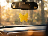 Yellow butterfly-shaped freshie hanging from a car rearview mirror with autumn trees through the windshield