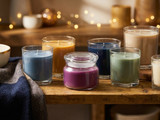 Seven colored glass-jar candles in blue, gold, navy, sage, cream, and a purple lidded jar on rustic wood with fairy lights