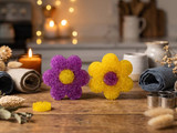 Purple and yellow flower-shaped aroma-bead freshies on a wood counter with a lit candle, dried flowers, and rolled towels