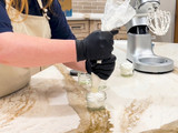 Black-gloved hands piping whipped white foaming bath butter scrub from a piping bag into a small glass jar on a marble counter, beside a stand mixer with a whisk attachment