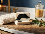 Two cream soap bars with black charcoal moustache embeds on a wood vanity beside a towel, sage, and candle