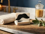 Two cream soap bars with black charcoal moustache embeds on a wood vanity beside a towel, sage, and candle