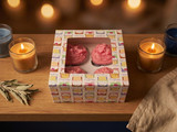 Pink whipped-wax cupcakes in a cupcake-print bakery box flanked by lit candles on wood