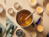 Overhead steel pitcher of amber melted wax with wood stir stick, ringed by candles, dropper bottles, dish of botanicals