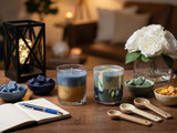 Two layered candles in glass jars on a wood table with notebook, pen, wax chips, measuring spoons, white roses, lantern