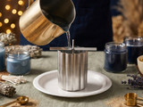 Melted wax pouring from a gold pitcher into a fluted metal mold on a paper plate, blue candles around