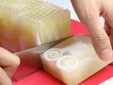 Hands slicing a bar from a translucent honey-toned melt-and-pour soap loaf on a red cutting board, the cut face showing two white spiral swirls and a bubble-textured top