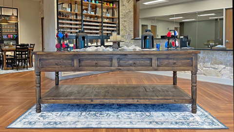 Rustic wooden display table on a blue patterned rug in a showroom, shelves of fragrance bottles and candle vessels behind