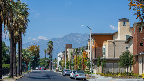 Palm-lined city street with parked cars and apartment buildings beneath hazy mountains under a blue sky
