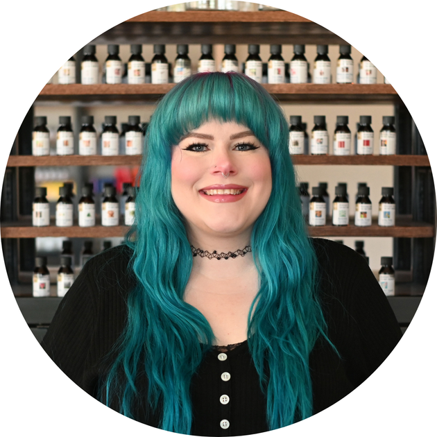 smiling woman with long teal hair and a choker in a black cardigan before shelves of fragrance bottles