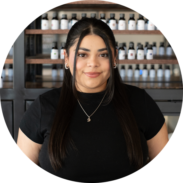 smiling woman with long straight dark hair and a pendant necklace before shelves of white bottles