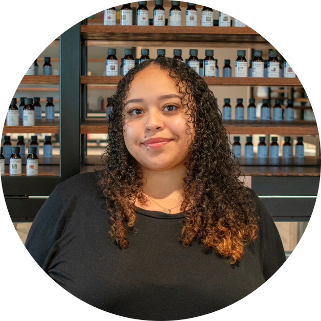 smiling woman with curly dark hair in a black top before wooden shelves of labeled bottles