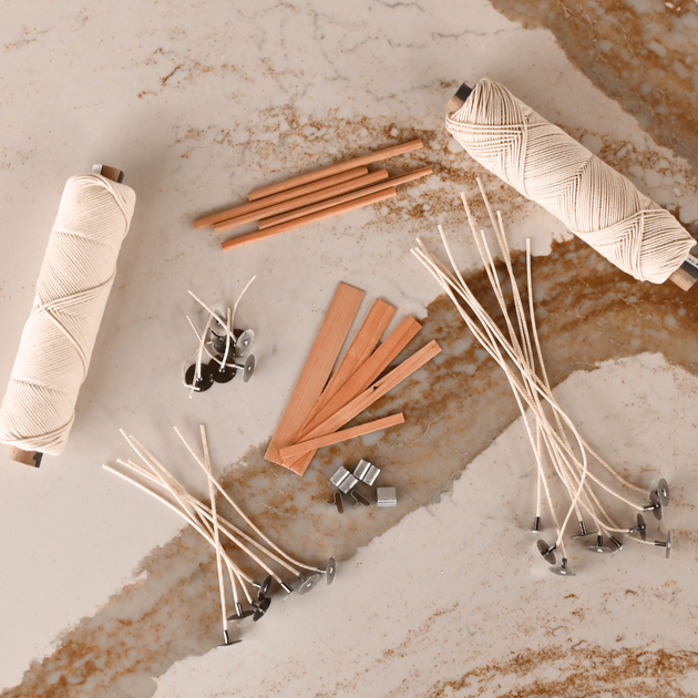 Assorted candle wicks on a marble surface: two spools of cotton wick, wooden wicks, and pre-tabbed waxed wicks with metal tabs