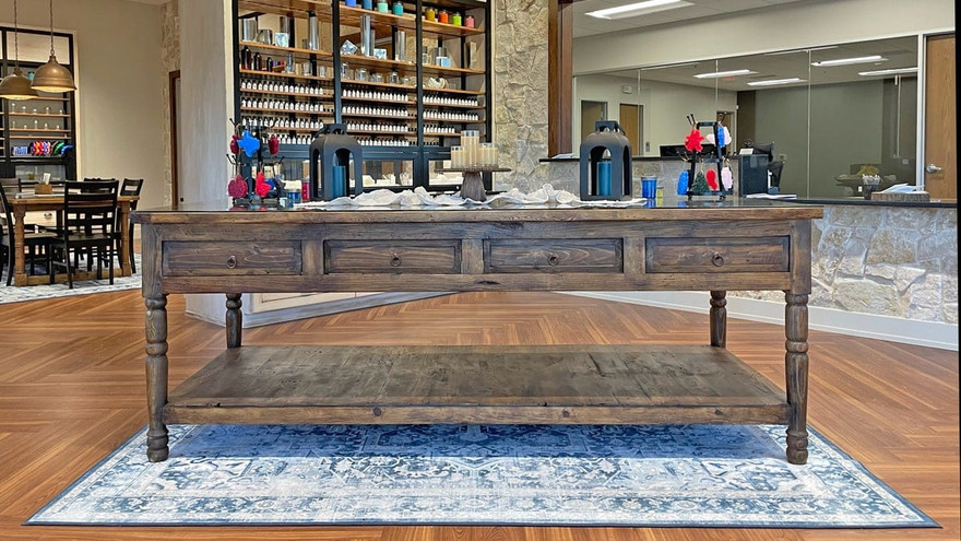 Rustic wooden display table on a blue patterned rug in a showroom, shelves of fragrance bottles and candle vessels behind