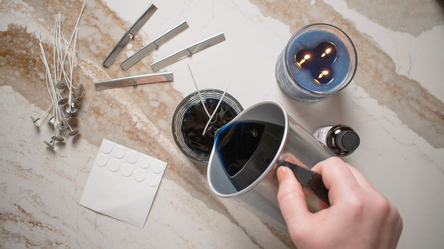 Overhead of hands pouring blue candle wax with wick pins, stickers, and a finished blue candle