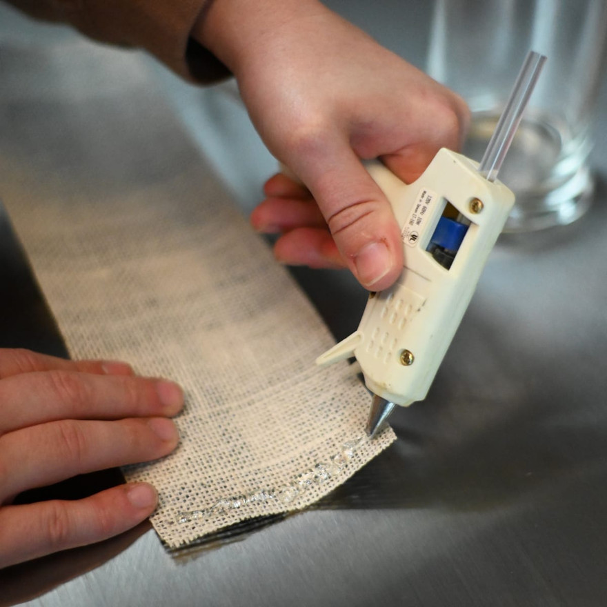 Hand running a hot glue gun along the end of a woven plaid ribbon strip on a metal counter