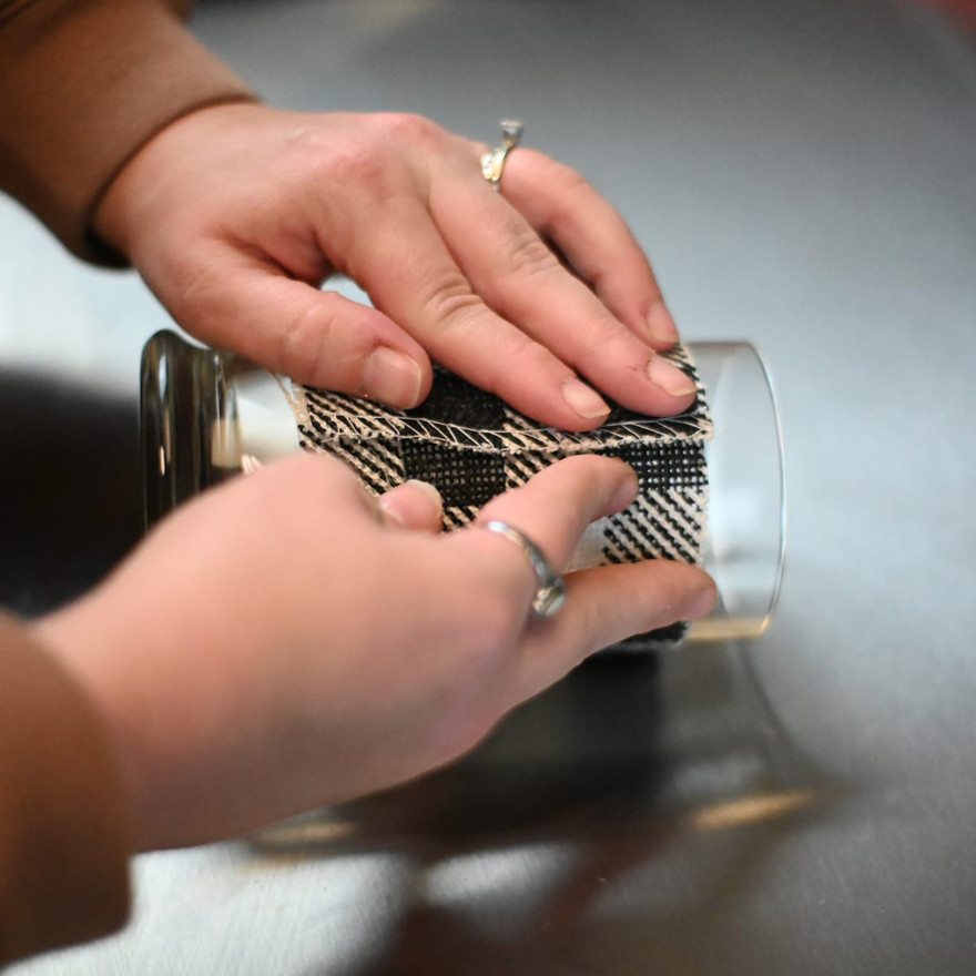 Hands wrapping black-and-white plaid ribbon around a clear glass jar laid on its side