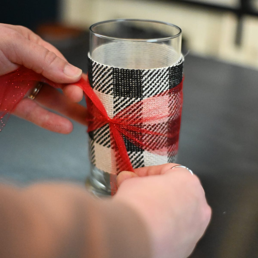 Hands wrapping red tulle around a clear glass wrapped in red-and-black plaid ribbon