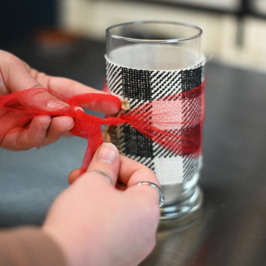 Hands tying red tulle into a bow over plaid ribbon on a glass jar, a wood tag tucked under