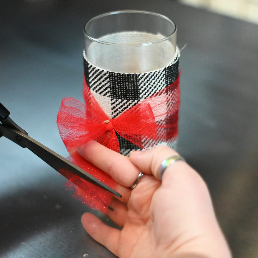 Black scissors trimming the tail of a red tulle bow on a plaid-wrapped glass jar