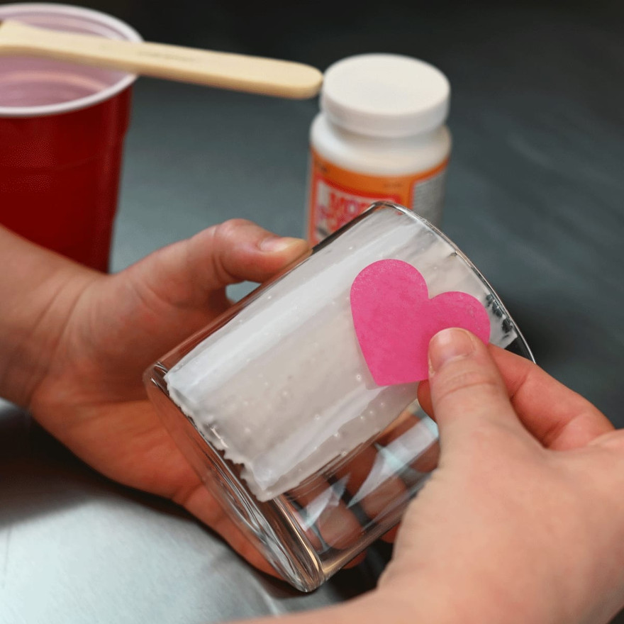 finger pressing a pink tissue paper heart onto a glue-coated glass jar