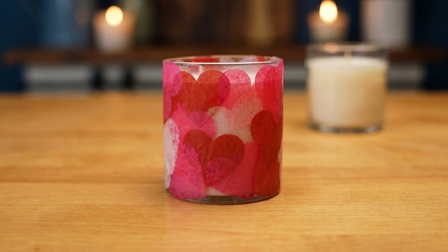 Glass candle vessel covered in overlapping red and pink tissue-paper hearts on a wood table beside a lit white candle