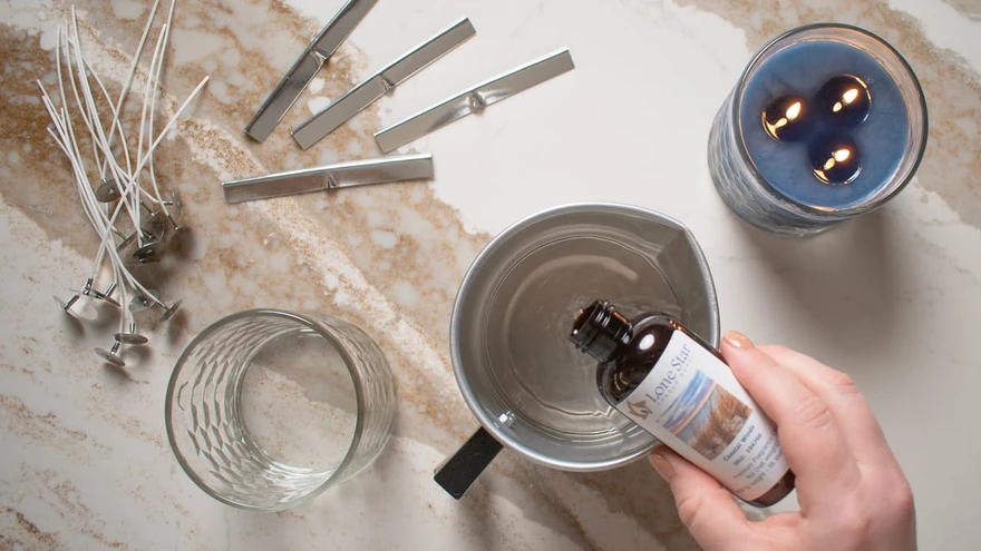 Overhead shot of a hand pouring a Lone Star fragrance oil bottle into a metal pour pot beside wicks, an empty jar, and a finished blue candle
