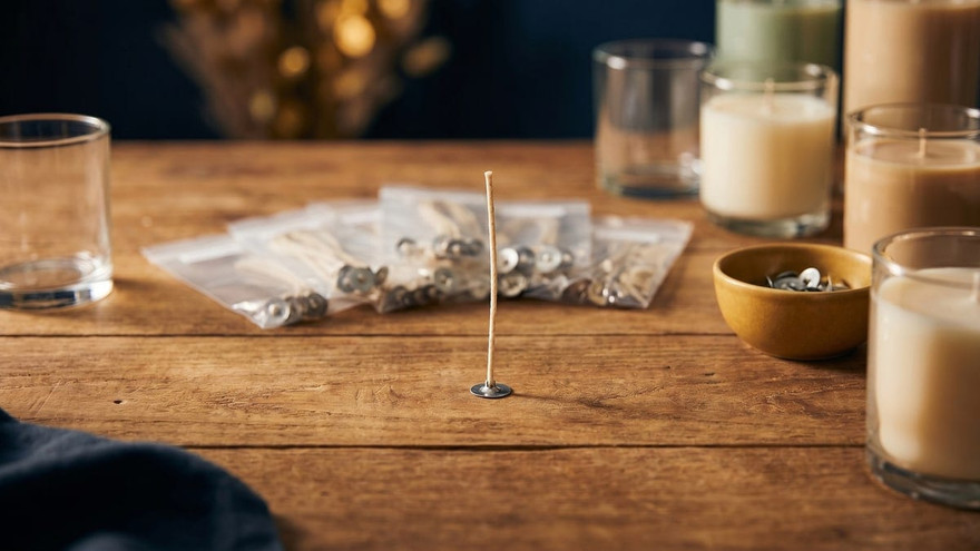 Single tabbed cotton wick standing upright on a wood table, bagged wick assortments and finished candles behind