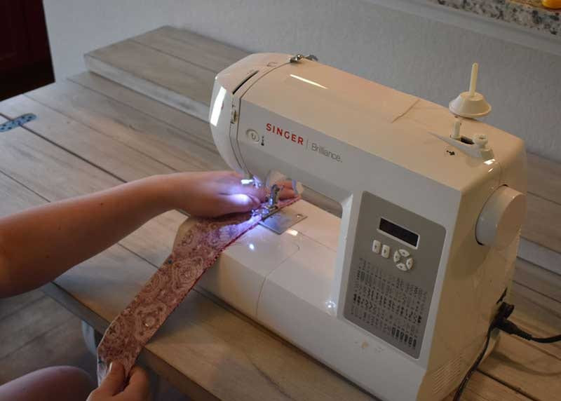 Hands feeding the pinned fabric strip through a Singer Brilliance sewing machine to stitch the seam