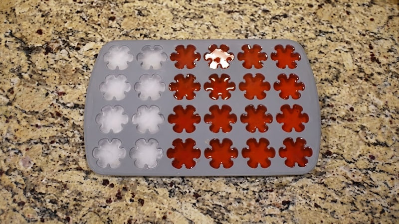 Gray flower silicone mold half-filled with melted red wax beside empty white cavities on granite counter