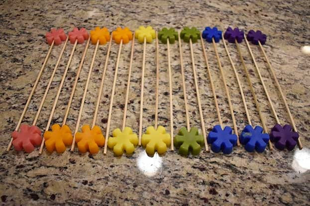 Row of red, orange, yellow, green, blue, and purple flower wax tarts mounted on wooden skewers on granite