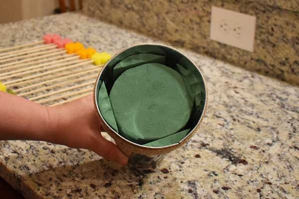 Hand holding a galvanized cup with a green floral foam disc fitted inside, by a counter outlet