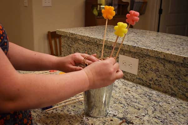 Hands arranging yellow and pink skewered wax tart flowers into a galvanized cup on a counter