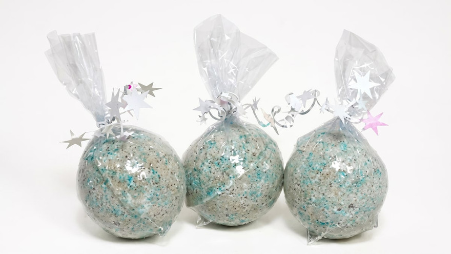Three round bath bombs speckled gray with teal flecks, wrapped in clear cellophane tied with silver star-spangled ribbon, on a white background