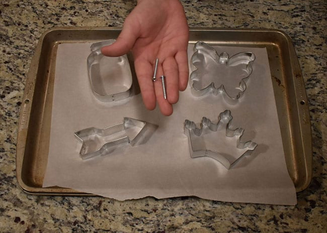 Hand holding two small metal pins above cookie cutters on a parchment-lined baking sheet