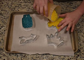 Pouring yellow aroma beads into a butterfly cookie cutter, blue-filled mason jar cutter beside it on a sheet