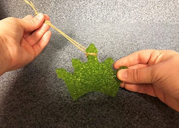 Hands pulling a twine hanger through a green crown-shaped beaded freshie on gray surface