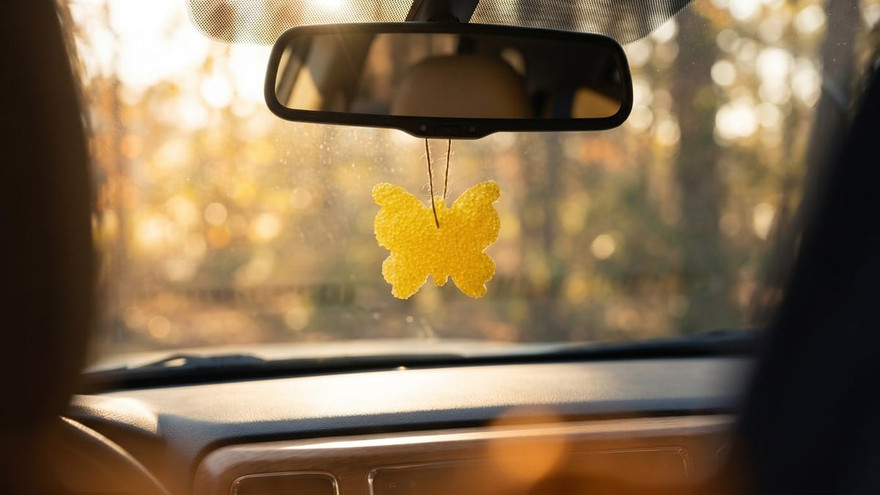 Yellow butterfly-shaped freshie hanging from a car rearview mirror with autumn trees through the windshield